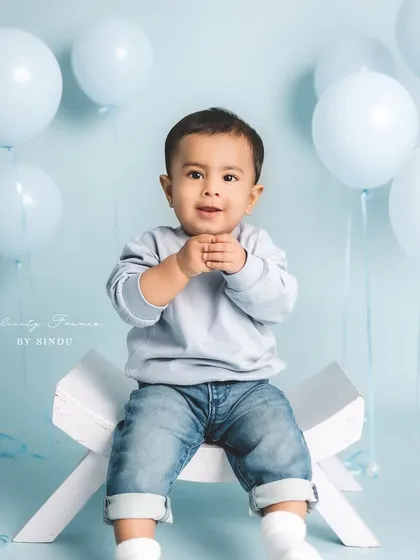 A handsome little man in jeans and a sweater, sitting confidently for his milestone portrait. The simple blue background makes his happy expression stand out.