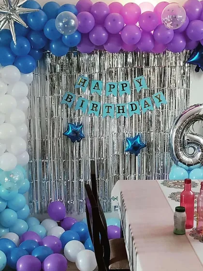 A fun and vibrant home decoration for a 6th birthday. This setup includes a multi-colored balloon arch, a silver foil fringe curtain, and star-shaped foil balloons to make the space feel festive.