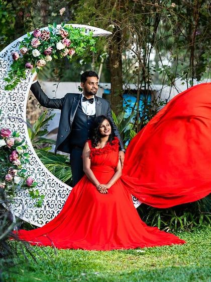 A romantic pose in a lush garden setting, with a large crescent moon prop and the bride's red gown creating a whimsical and dreamy pre-wedding photo.