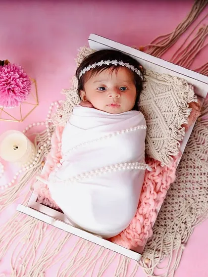 A newborn wrapped in a simple white swaddle with pearl details, resting in a miniature bed. The pink background and macrame textures create a lovely boho-chic vibe.