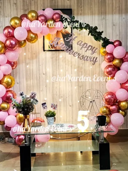 A lovely anniversary setup featuring a half-ring of pink and chrome gold balloons. The table is set with photos and props to celebrate the couple's journey.