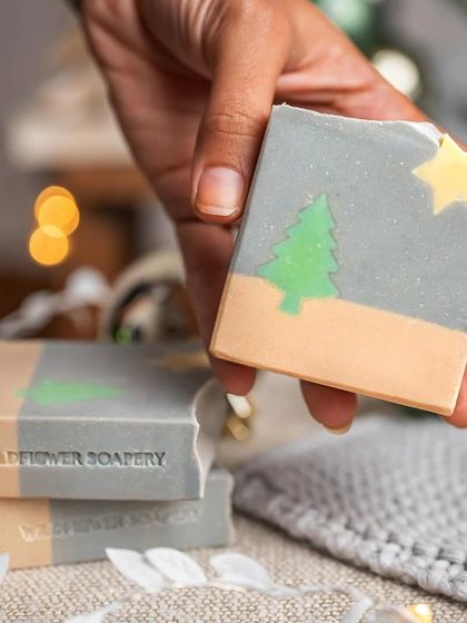 A hand holding a soap bar decorated with a simple tree and star. The soft, warm lighting from the fairy lights adds to the cozy holiday feeling.