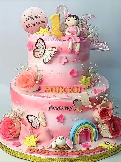 A beautiful two-tier doll-themed cake for a first birthday. The pink ombre design is adorned with a fondant doll, butterflies, a rainbow, and real roses.