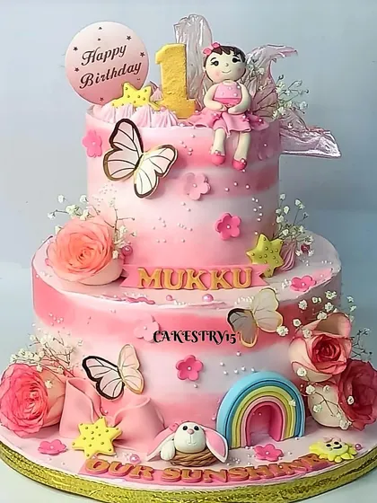 A beautiful two-tier doll-themed cake for a first birthday. The pink ombre design is adorned with a fondant doll, butterflies, a rainbow, and real roses.