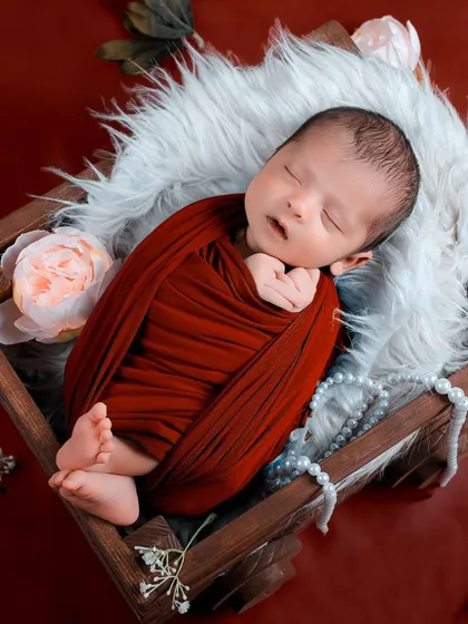 A baby wrapped in rich red fabric sleeps in a rustic wooden crate lined with soft fur and pearls, combining classic and rustic elements.