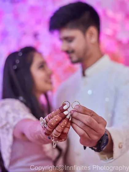 A beautifully lit shot focusing on the couple's hands holding their rings, with a soft pink floral background. The focus is on the rings as a symbol of their union.