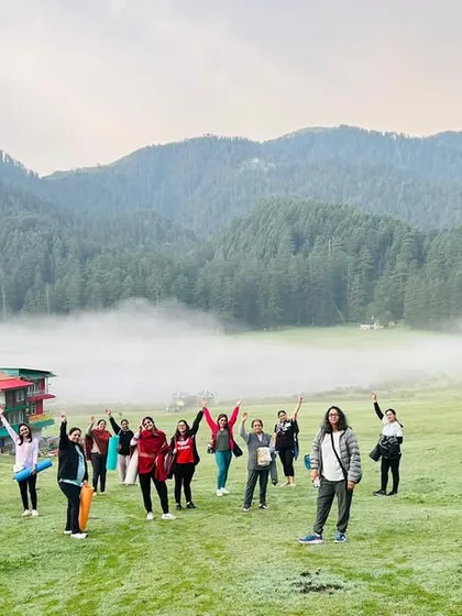 A wide view of our group celebrating the morning with arms raised, feeling the expansive energy of the Himalayan landscape. The background shows the dense deodar forests that circle the Khajjiyar valley.