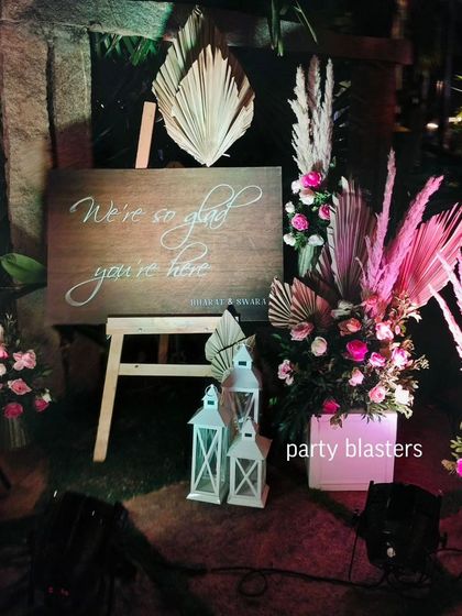 A rustic wooden welcome sign for a wedding, surrounded by pampas grass and pink floral arrangements. The sign reads, "We're so glad you're here," creating a warm and inviting entrance.