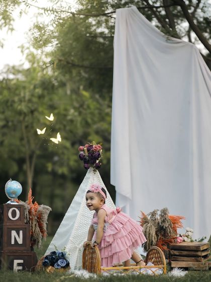 A little girl in a lovely pink dress explores her first birthday setup in a park. This candid shot captures her curiosity and movement, with a teepee and "ONE" blocks creating a magical, rustic scene.