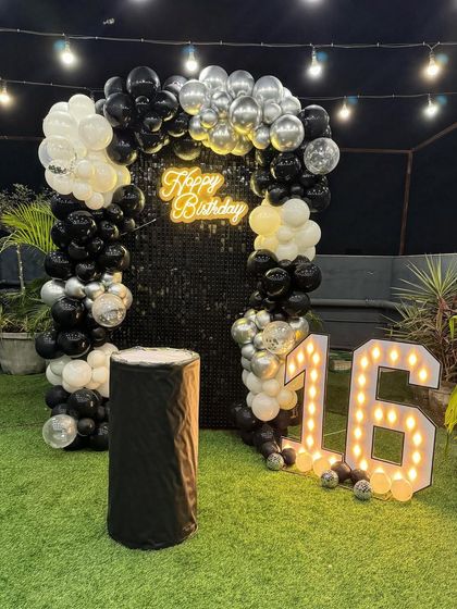 A sleek and modern 16th birthday setup on a terrace with an artificial grass floor. The black sequin shimmer wall is framed by a black, white, and silver balloon arch, complemented by large marquee numbers.