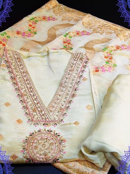 A gorgeous off-white unstitched suit set with a V-neckline featuring pink and gold embroidery. The set is completed with a stunning Banarasi Bandhej dupatta.