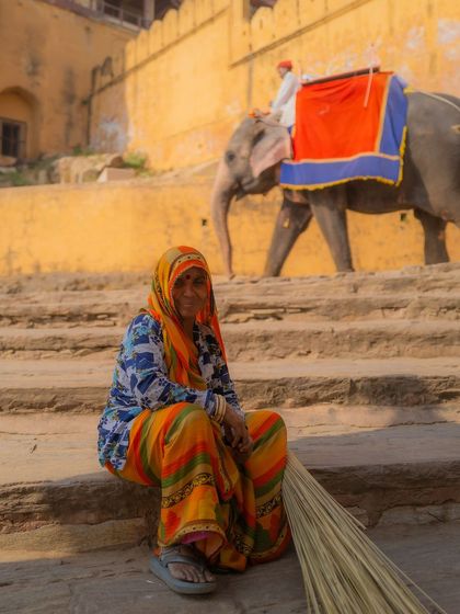 A woman in a colorful saree takes a break from sweeping the steps at Amer Fort in Jaipur. In the background, a decorated elephant walks by, creating a classic Rajasthani scene.
