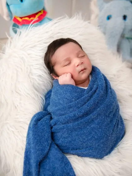 A newborn is swaddled in a cozy blue wrap, resting on a white fur blanket with plush toys in the background. This image shows a simple yet sweet setup, focusing on the baby's serene sleep.