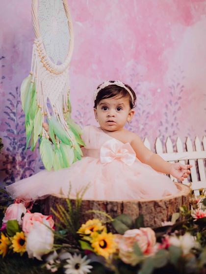 A portrait of a baby girl in a beautiful pink dress, seated on a tree stump prop with a dreamcatcher in the background. The floral and bohemian elements create a unique, artistic look.