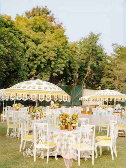 A classic daytime garden party setup. The use of traditional printed umbrellas, white furniture, and yellow floral centerpieces creates a cheerful and elegant atmosphere for a corporate brunch or luncheon.