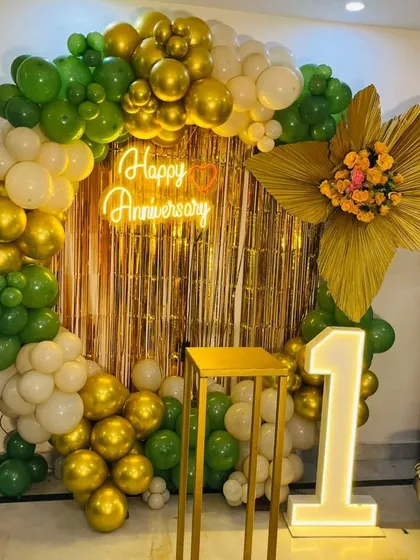 A first-anniversary decoration with a gold fringe curtain. The setup features a circular balloon arch in green, white, and gold, a neon sign, and a marquee number '1'.