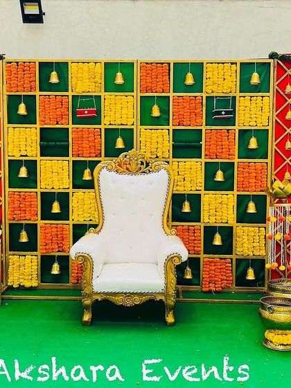 A classic Seemantham stage design with a colorful checkered backdrop made of woven fabric. The setup is accented with traditional brass bells and framed by red lattice panels.