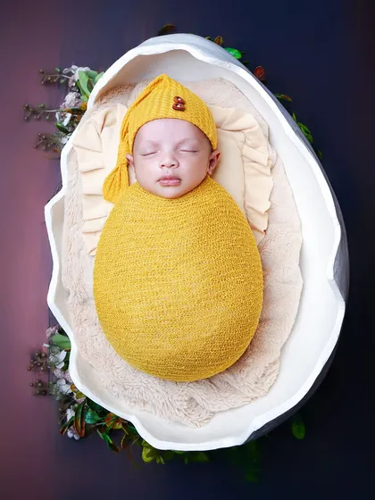 A close-up shot of the baby nestled in the eggshell prop, wearing a cozy yellow hat. This highlights the baby's peaceful expression within the creative setup.