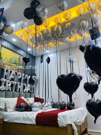 This view shows the "Happy Birthday" foil balloon banner against a dark wall, complemented by black and silver balloons on the ceiling and bed.