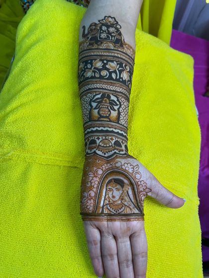 A detailed view of the arm portion of a bridal mehndi. This section features a bride's portrait, an elephant motif, and a sacred kalash, all drawn with precision.