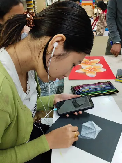 A student using her phone for reference while working on a detailed drawing with pastels, blending modern tools with traditional techniques.