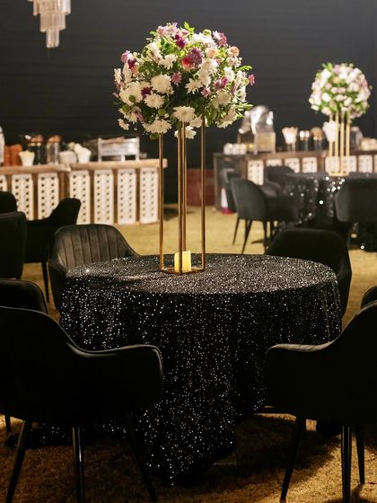 A glamorous table setting for a Sangeet, with a black sequined tablecloth, a tall floral centerpiece, and elegant black chairs.