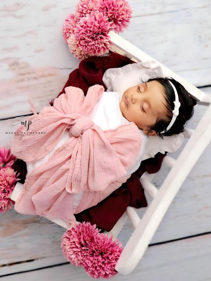 A baby girl wrapped in a pink bow, resting on a white diwan with pink pom-pom flowers.