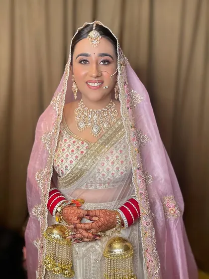 A full view of a bride in her beautiful white and pink lehenga. The look is completed with a stunning polki necklace, maang tikka, and traditional red bridal bangles.