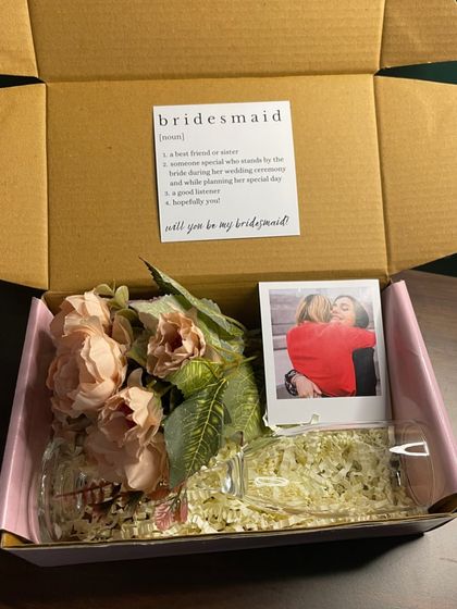 A beautiful bridesmaid proposal box featuring a mini champagne glass, a small flower bouquet, a polaroid, and a definition card. It’s an elegant and classy way to pop the question to your friends.