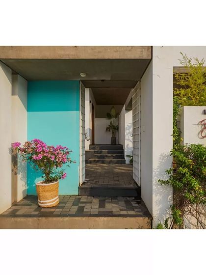 The entrance to the Aayi House features a bright pop of turquoise on the door, offering a playful contrast to the otherwise minimalist white and concrete structure. The name 'Aayi' is elegantly scripted in Kannada.