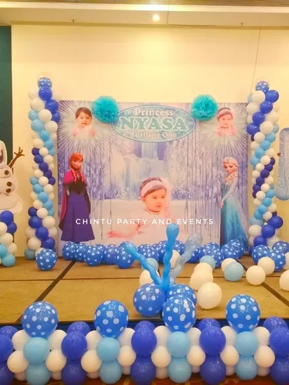 A Frozen themed first birthday party stage. The setup features a winter-themed backdrop with characters, framed by blue and white balloon pillars.