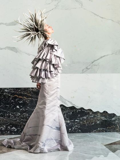 A profile shot that emphasizes the avant-garde headpiece and the texture of the gown against a backdrop of black and white marble. The lighting is sculpted to highlight the model's form and the dramatic styling.