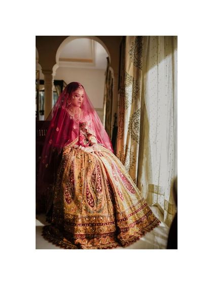 A classic bridal portrait. The bride sits gracefully by a window, her intricate lehenga spread out, creating a regal and timeless image.
