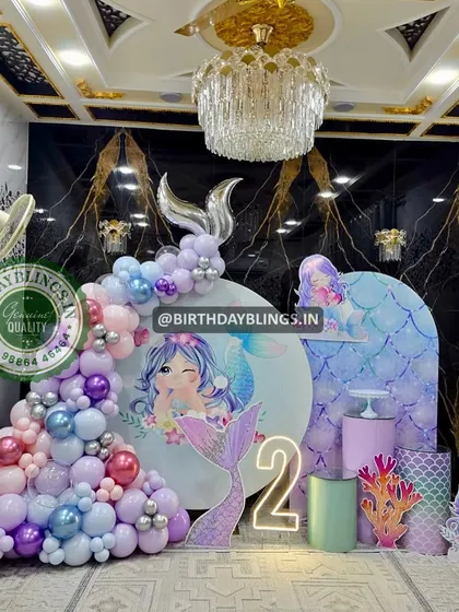 A glamorous mermaid-themed second birthday party set against a marble wall. The decor features iridescent and pastel balloons, a neon number '2', and shimmering mermaid tail cutouts.