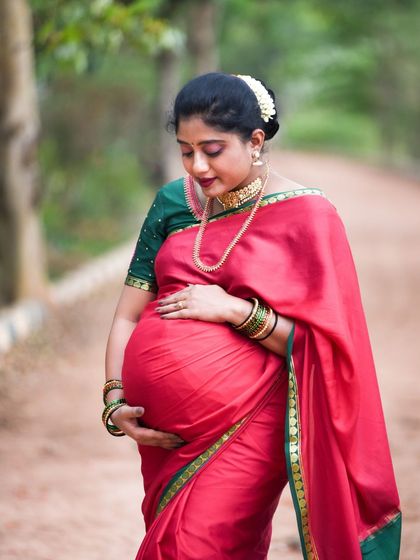 An elegant outdoor portrait of a mom-to-be in a traditional red saree, capturing a moment of quiet reflection.