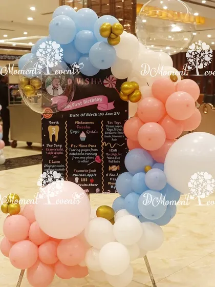 A Krishna-themed milestone board for a first birthday. The board is framed by a circular arrangement of pink, blue, and white balloons with gold accents.