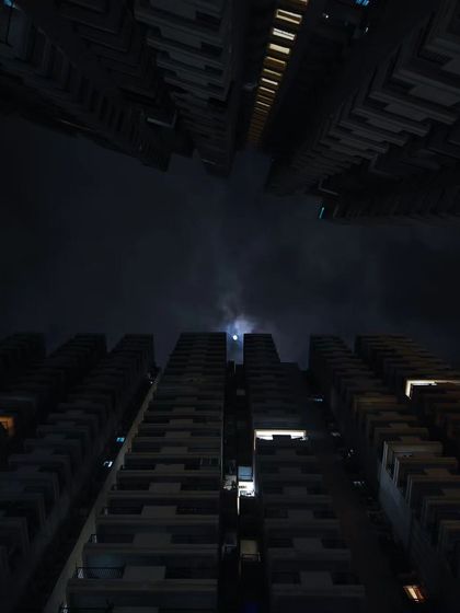 A similar low-angle shot of city buildings, but at night. The composition creates a moody, almost dystopian feel, framed by the dark silhouettes of the architecture.