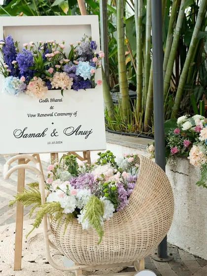 A beautiful welcome sign for a Godh Bharai and vow renewal ceremony. The sign is adorned with a shadow box filled with pastel flowers, and a wicker basket overflowing with similar blooms sits at its base.