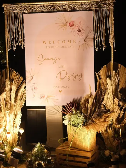 A personalized welcome sign for a boho-chic cocktail party. This setup combines elegant calligraphy with natural elements like pampas grass, dried palms, and a macrame frame to set a warm and stylish tone right from the entrance.