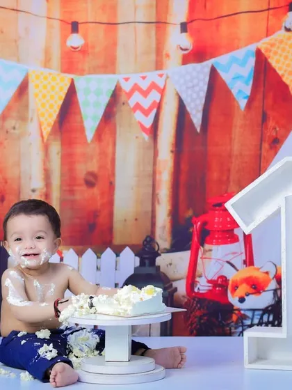A classic cake smash scene. A one-year-old boy sits in front of the number "1" with cake all over his face and hands, smiling happily.