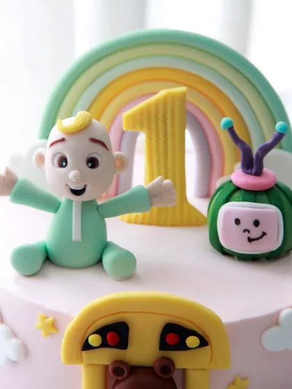 A close-up of the adorable, handcrafted fondant toppers on the Cocomelon cake, including JJ and the smiling melon under a pastel rainbow.