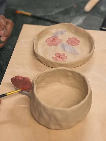 A student painting delicate pink flowers onto a raw clay bowl. This is underglazing, done before the final firing.