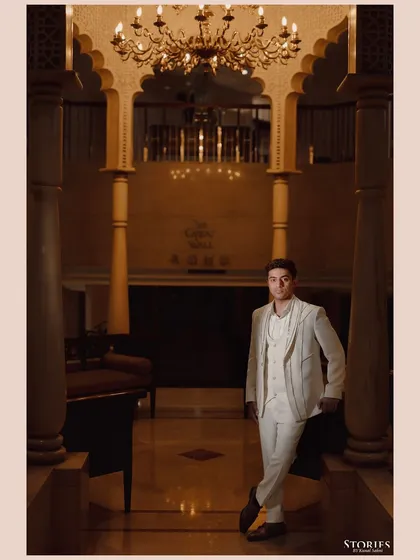 A stylish portrait of the groom, Rupen, in his all-white engagement suit, standing in a grand, beautifully lit hall.