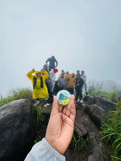 A creative shot holding up our trek badge, with the happy group posing on the rocks at the Kurinjal peak in the background.