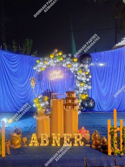 An outdoor birthday stage at night, with a blue curtain backdrop and a silver and gold balloon arch.