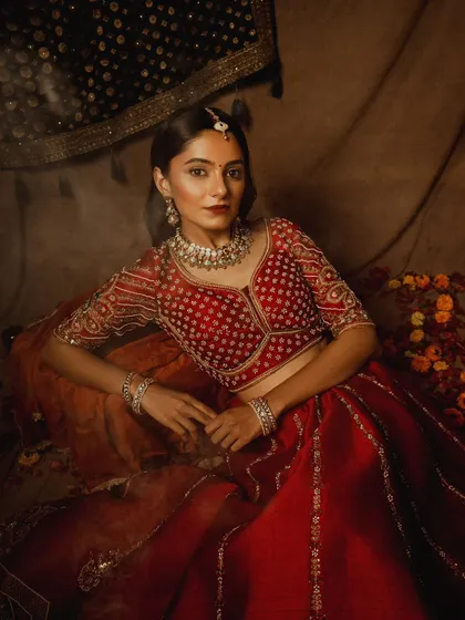 A classic bridal portrait, with the model seated amongst flowers. The lighting is soft and flattering, highlighting the rich red of the lehenga and the sparkle of the jewelry.