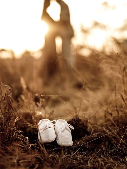 An artistic and romantic announcement. The focus is on the tiny baby shoes in the foreground, with the couple sharing a kiss in the beautifully blurred, golden-hour background.