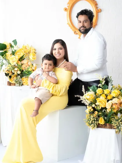 An elegant family portrait of three. The mother sits gracefully holding her son while the father stands beside them, creating a beautifully composed shot with a lovely yellow floral arrangement.