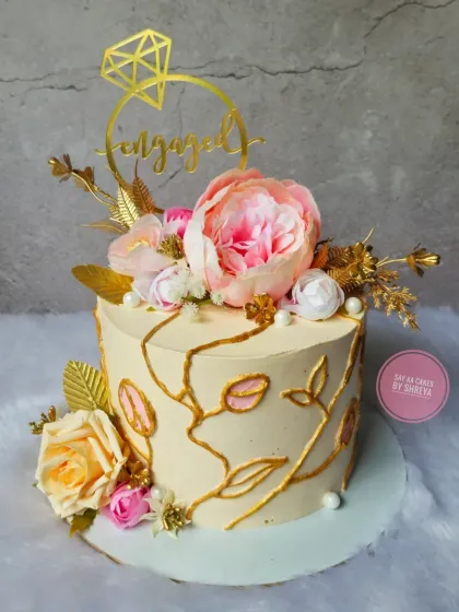 An elegant cake to announce an engagement. The design features beautiful pink and cream flowers, delicate gold vine details, and a "Engaged" ring topper, symbolizing a beautiful new beginning.