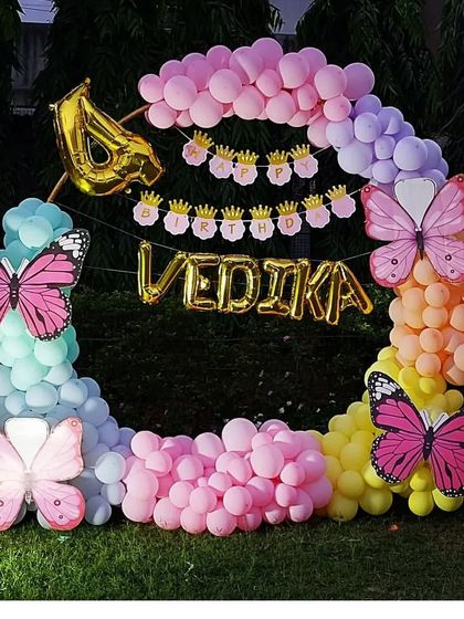 A colorful outdoor butterfly-themed party for a 4th birthday. The circular balloon arch in pastel colors is decorated with large butterfly cutouts and the birthday girl's name.